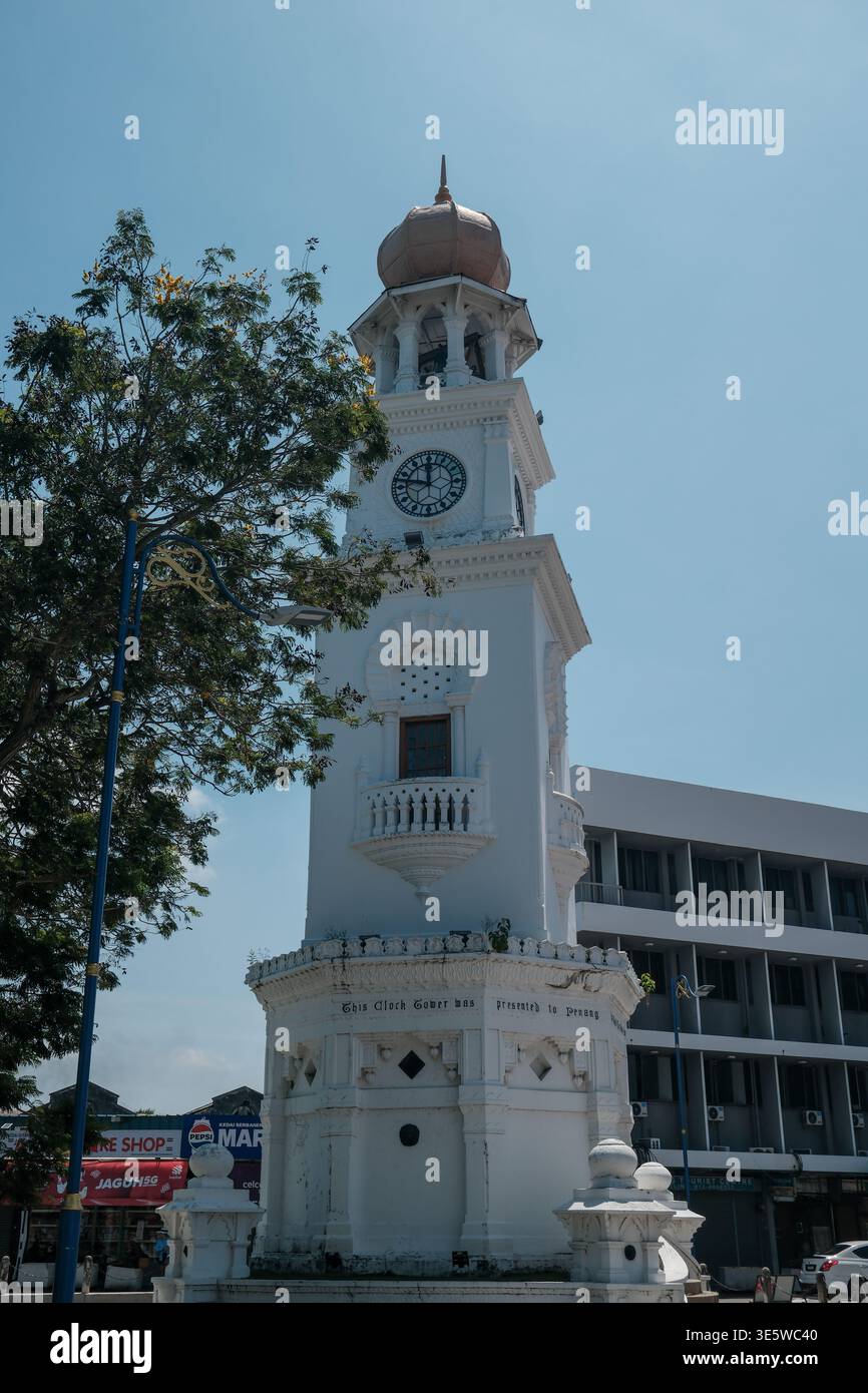 White clock tower stands tall against clear sky. Decorative arches frame its ornate clock face. Tree branches frame the tower's left side. Modern buil Stock Photo