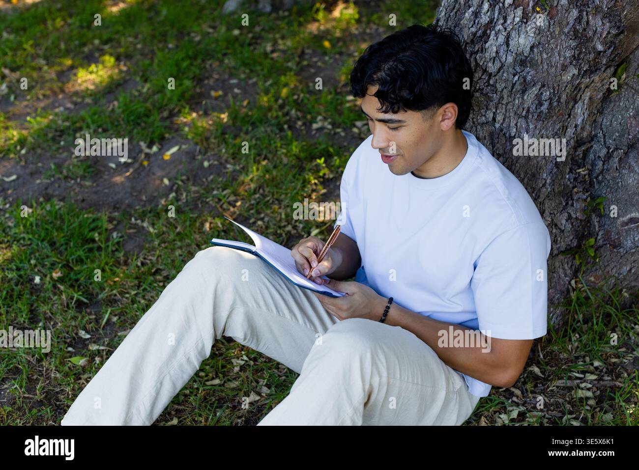 Asian man sitting on grass by trunk writing journal with pencil white-tee beige-pants, copy space Stock Photo