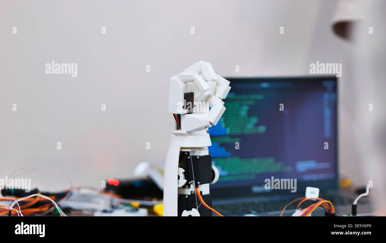 A robotic arm prototype on a desk with a laptop showing programming code in the background Stock Photo