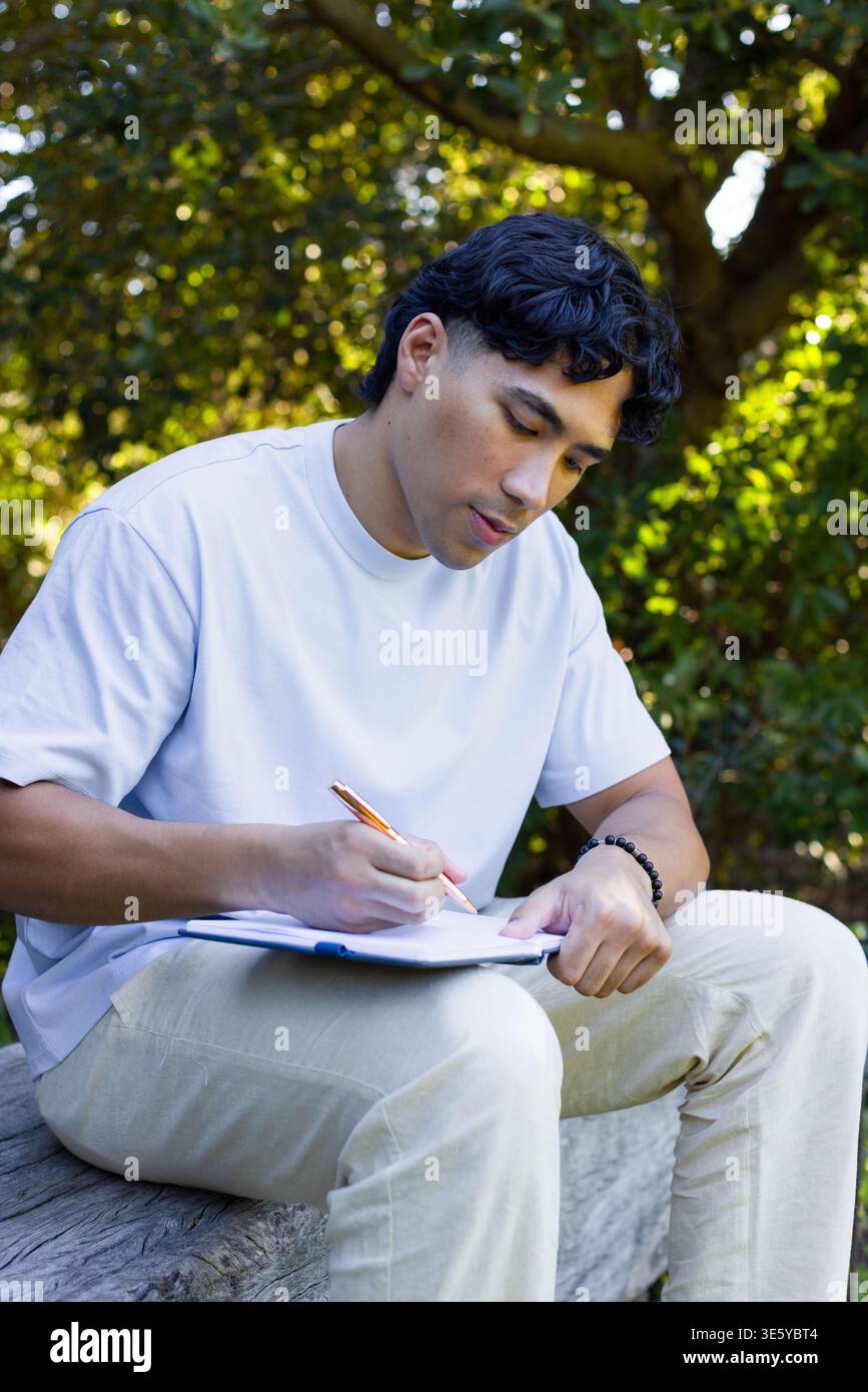 Adult male sitting on log at park writing on pad with orange-pen, white tee, beaded bracelet Stock Photo