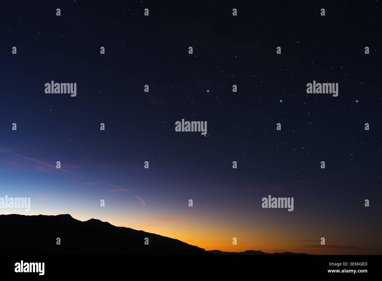 Early dawn light coloring the horizon orange above a silhouetted mountain range in the California desert, with stars still visible overhead. Stock Photo