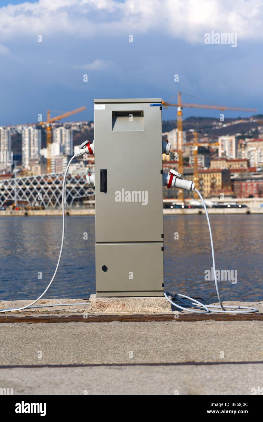 A shore power pedestal with multiple heavy-duty electrical connectors and cables located on a dockside. The background features calm water, high-rise Stock Photo