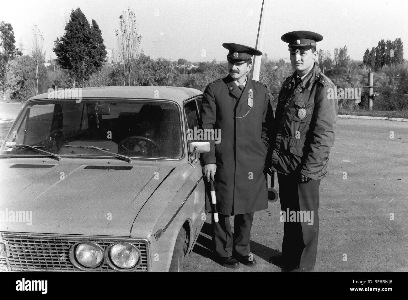 Slaviansk, 1997, on a city bridge road near the pencil factory. An archive black and white photo showing two traffic police officers during a vehicle inspection. The uniformed inspectors are standing next to a light-colored passenger car, holding a striped traffic wand and looking attentively to the right side of the frame. The famous monument with giant pencils, autumn trees, and tall electrical towers are visible in the background. A daily police routine. This is a scene of peaceful life in Donbas. Stock Photo