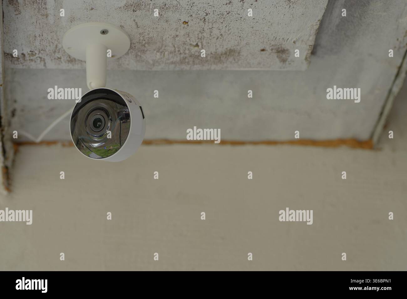White security camera mounted under a textured ceiling, positioned in the upper corner with visible bracket and reflective lens, set against neutral w Stock Photo
