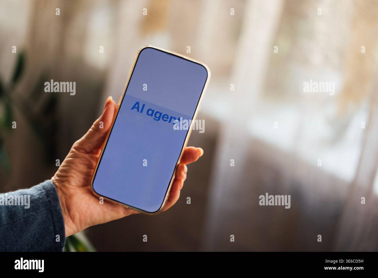 Hand of a person holds a smartphone showing the text "AI agent," with a soft-focus background of curtains and indoor plants in a cozy interior Stock Photo