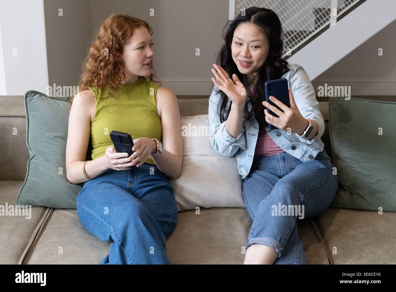 Two women sitting close on beige sofa, holding smartphones, one waving and wearing denim jacket Stock Photo