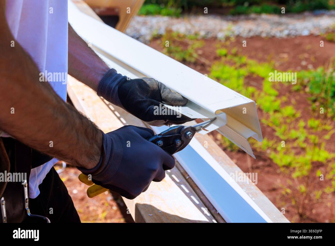Carpenter uses scissors to cut white piece of vinyl siding while working outdoors on construction project. Stock Photo