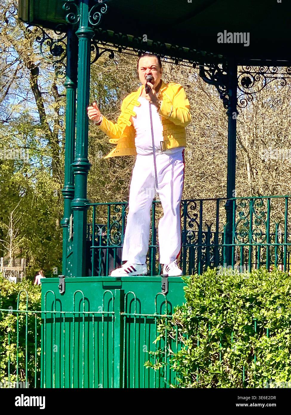 Freddie Mercury lookalike dressed in a white and red stage style costume poses in Abington Park Northampton near the bandstand confidently on a Sunday - Smartphone Captured Stock Image