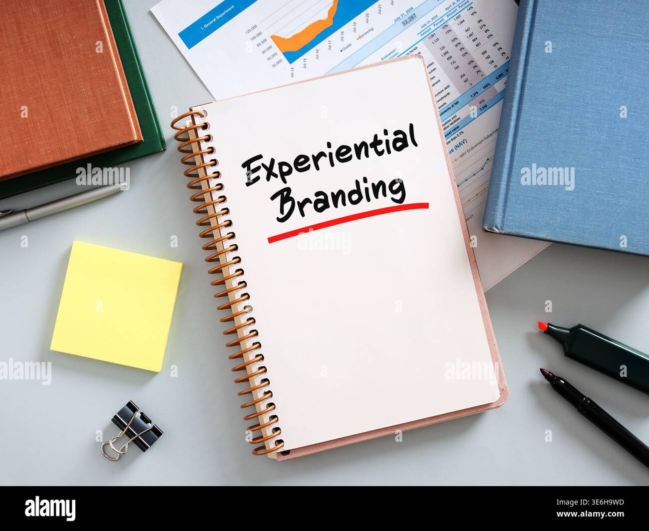 Notebook with experiential branding handwritten on it, placed on desk with pen, highlighter, documents, books, sticky notes and binder clip Stock Photo