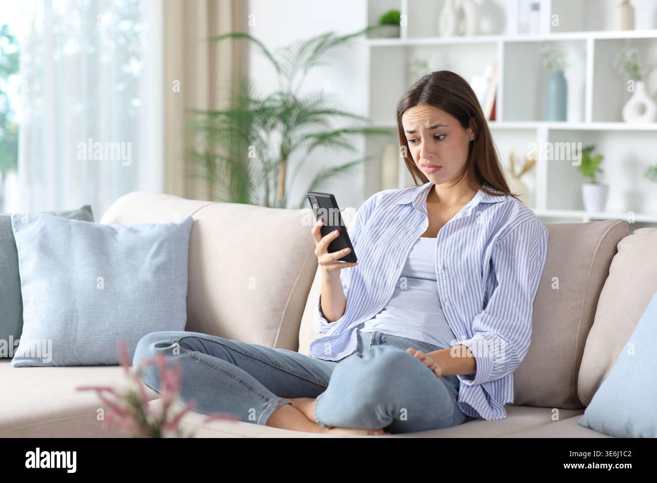 Disappointed woman in blue checking phone sitting on a couch at home Stock Photo