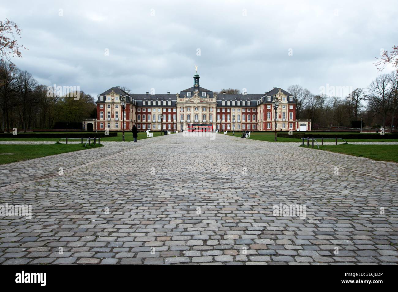 Muenster, Germany - March 30, 2025: Wide view of Schloss Muenster with cobblestone square and a red bus in front of the baroque building under cloudy Stock Photo