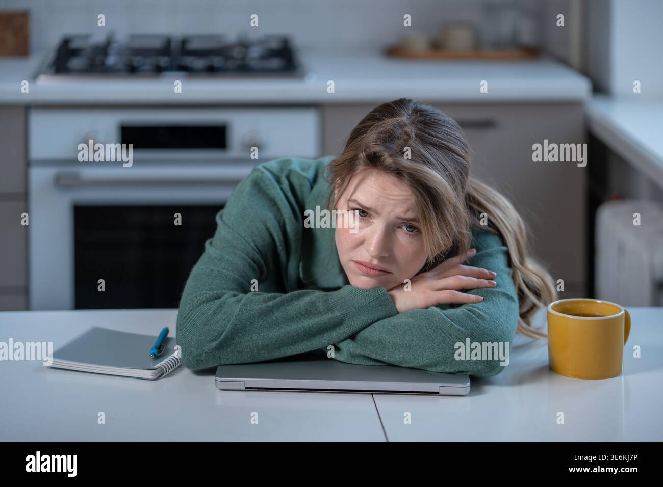Tired frowning woman lying on closed laptop at kitchen table near cup coffee. Burnout, low productivity while working from home. Exhausted young femal Stock Photo