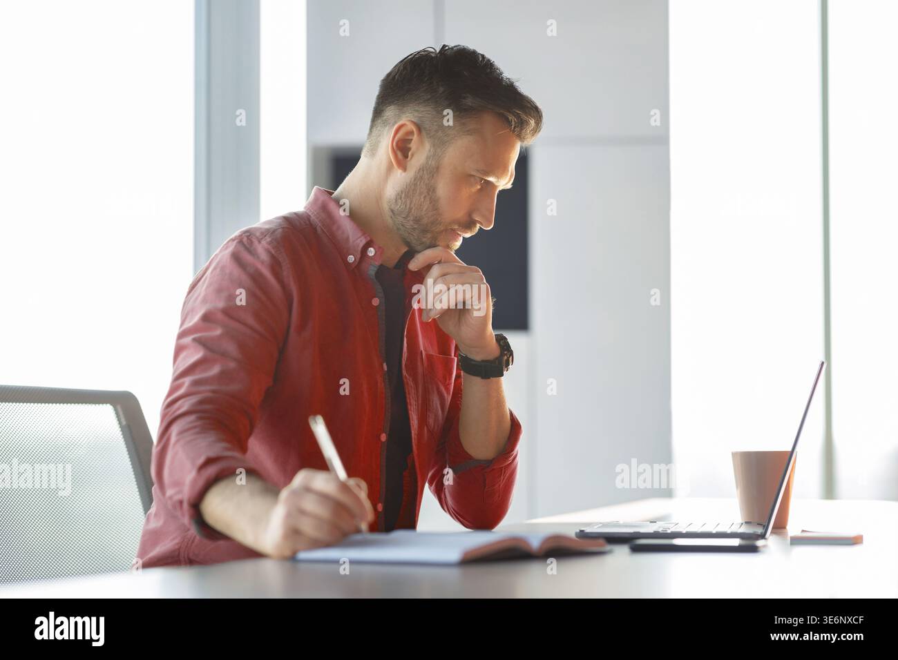 Man Writing Notes While Looking at Computer in a Bright Office Setting in the Afternoon Stock Photo