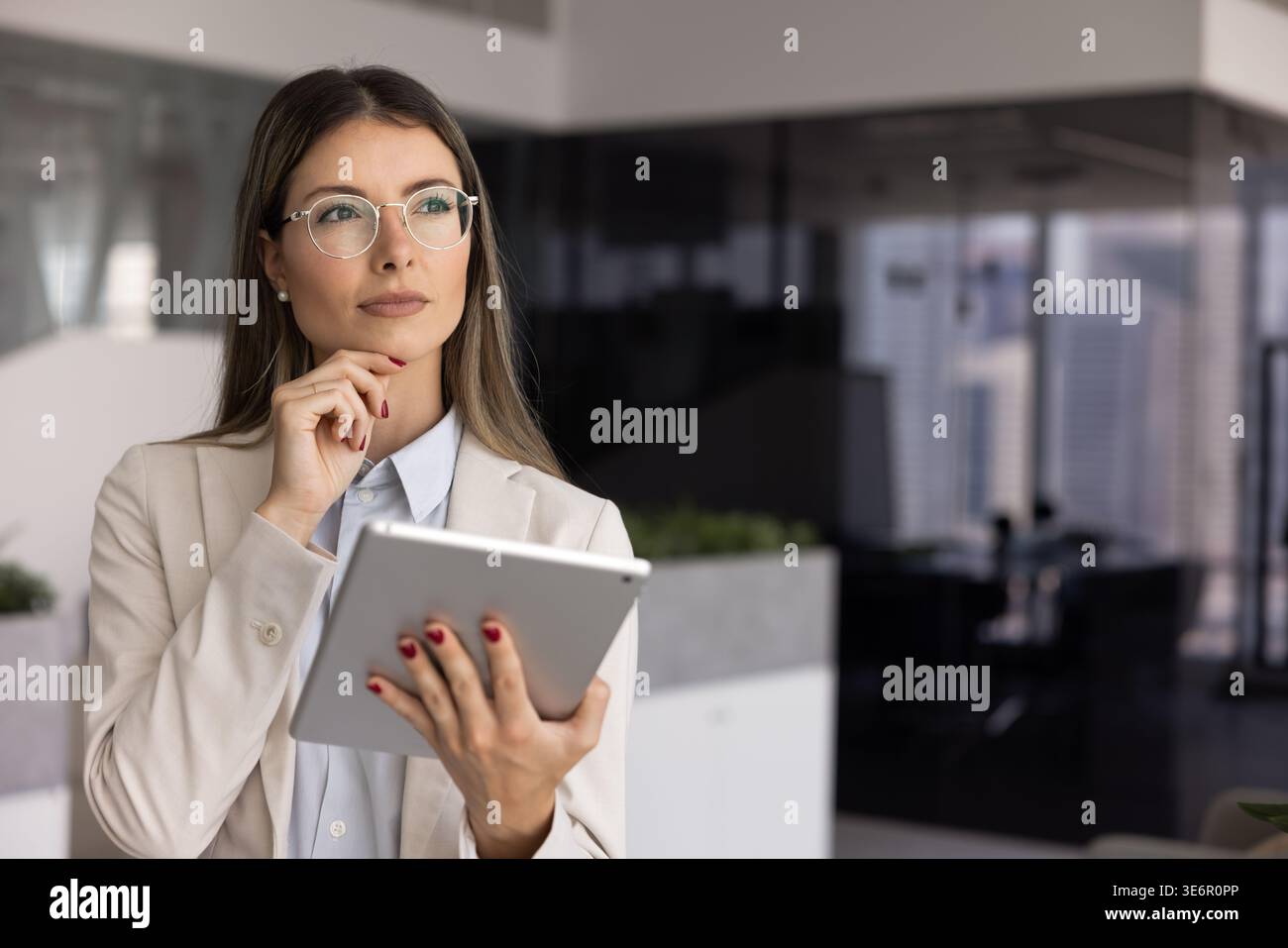 Thoughtful professional woman stands in modern office with tablet Stock Photo