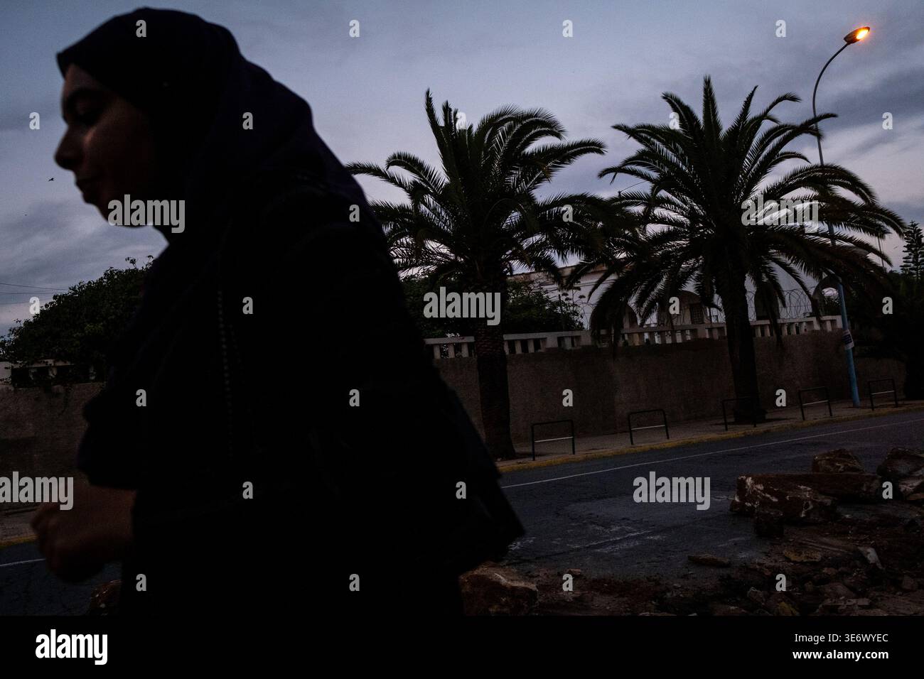 A pedestrian in dark clothing walks along a palm lined street at dusk in the medina of Casablanca, Casablanca-Settat, Morocco, October 18, 2023. Casab Stock Photo
