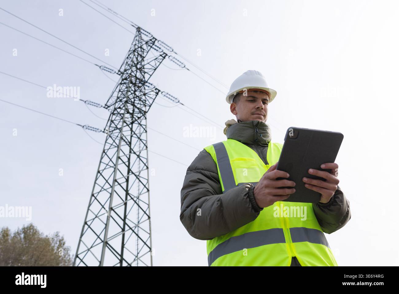 Young male engineer uses a tablet to check high-voltage power lines while working outdoors in energy infrastructure. Stock Photo