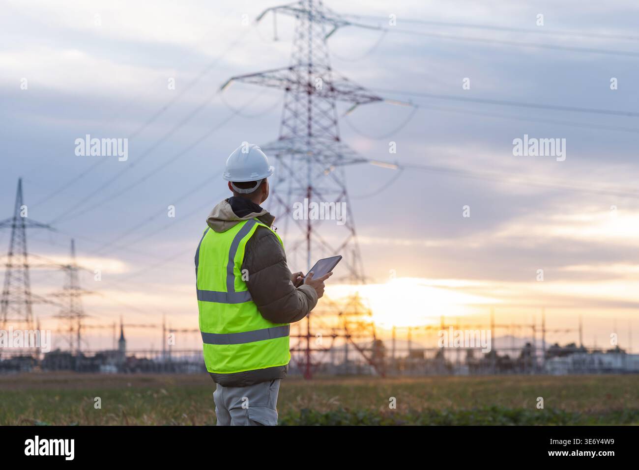 A young male engineer checks high-voltage power lines with a tablet outdoors during sunset, focusing on safety and energy management. Stock Photo