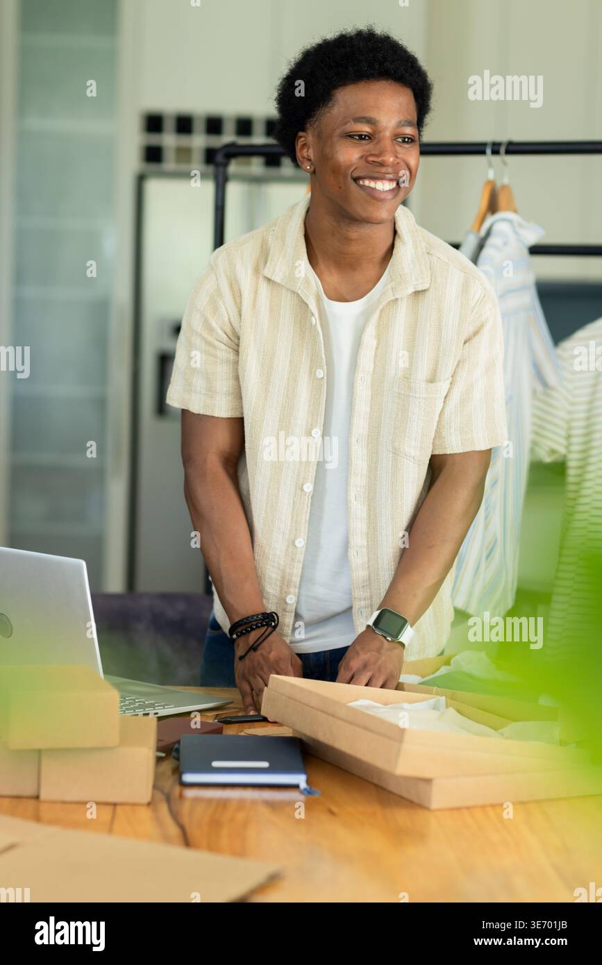 Man packing cardboard boxes on wooden table with laptop and clothing rack in small home studio Stock Photo
