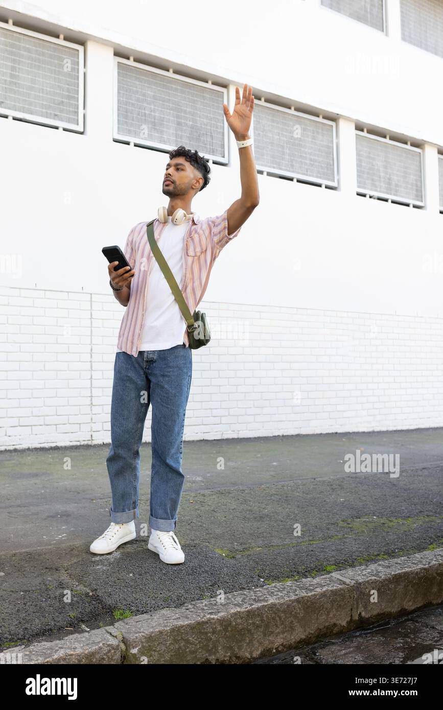 Man in 20s holding phone, raising hand on curb by white wall, wearing headphones and bag. Urban, streetwear, casual, crossbody, outdoors, daytime, ges Stock Photo