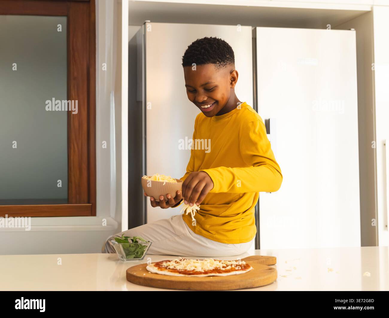 African American child wearing yellow top sprinkling cheese over pizza on countertop in kitchen. Kid, smile, prep, greens, spinach, bowl, modern Stock Photo