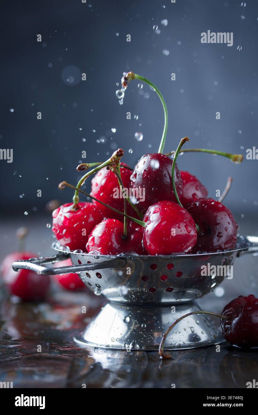 Fresh red cherries, plump and vibrant, are generously piled into a gleaming stainless steel colander. Water splashes are dynamically frozen in mid-air Stock Photo