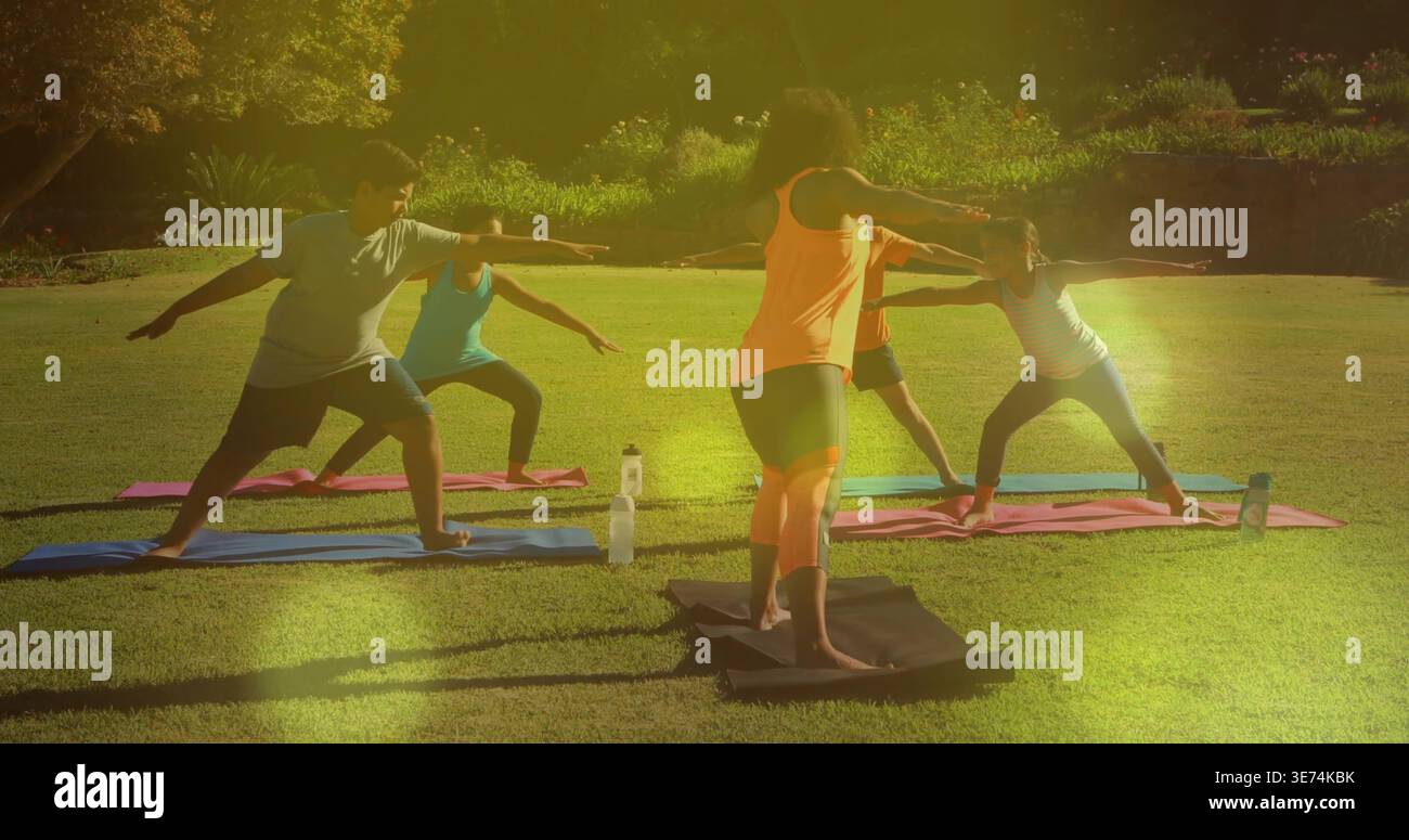 Leading instructor in orange tank guiding children doing Warrior II on mats in park, water bottles. Outdoor, yoga, leader, kids, group, sunshine, natu Stock Photo