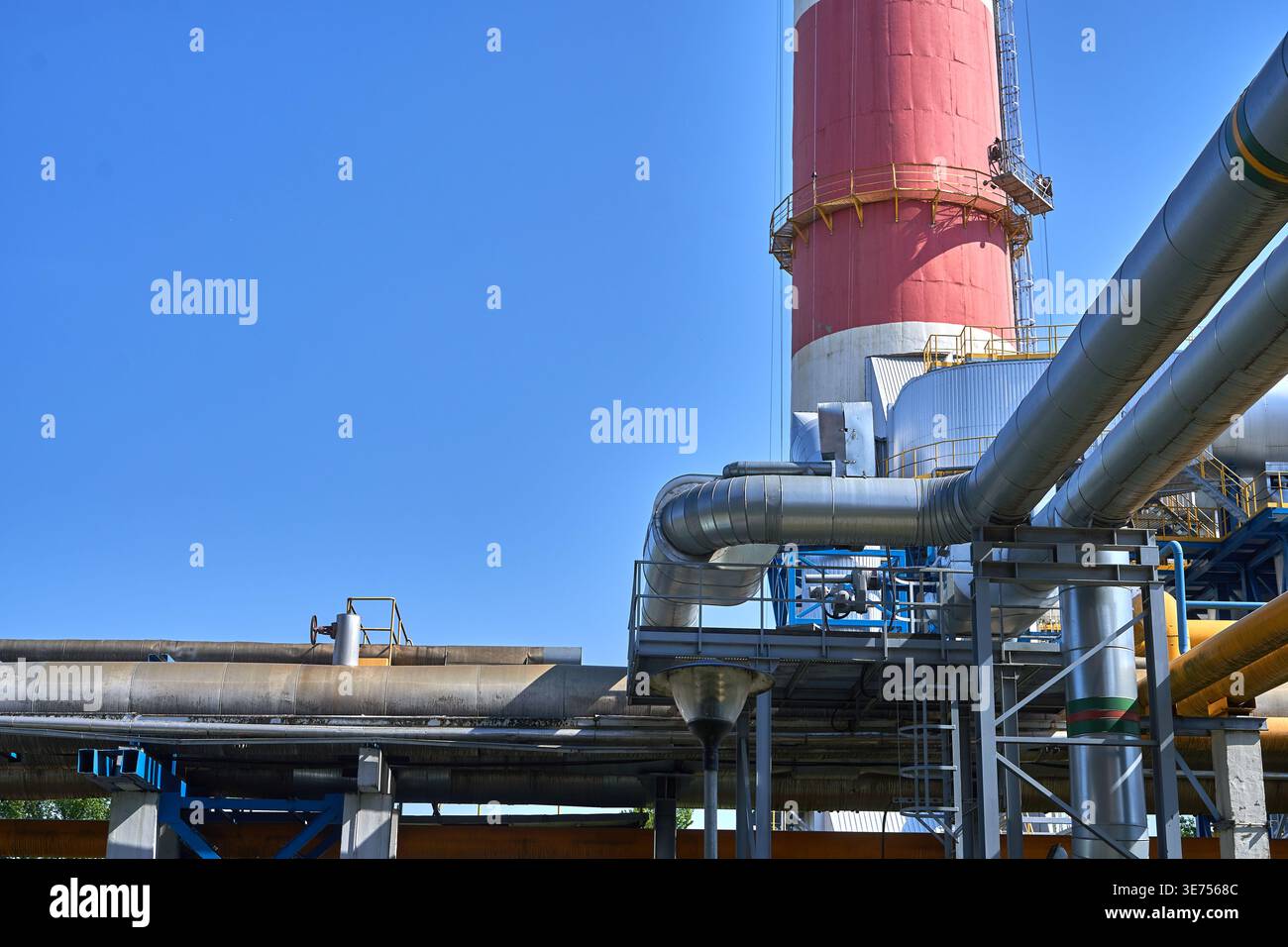 Industrial facility with extensive metal piping networks and a prominent red and white chimney stack against clear blue sky. Large silver pipelines Stock Photo
