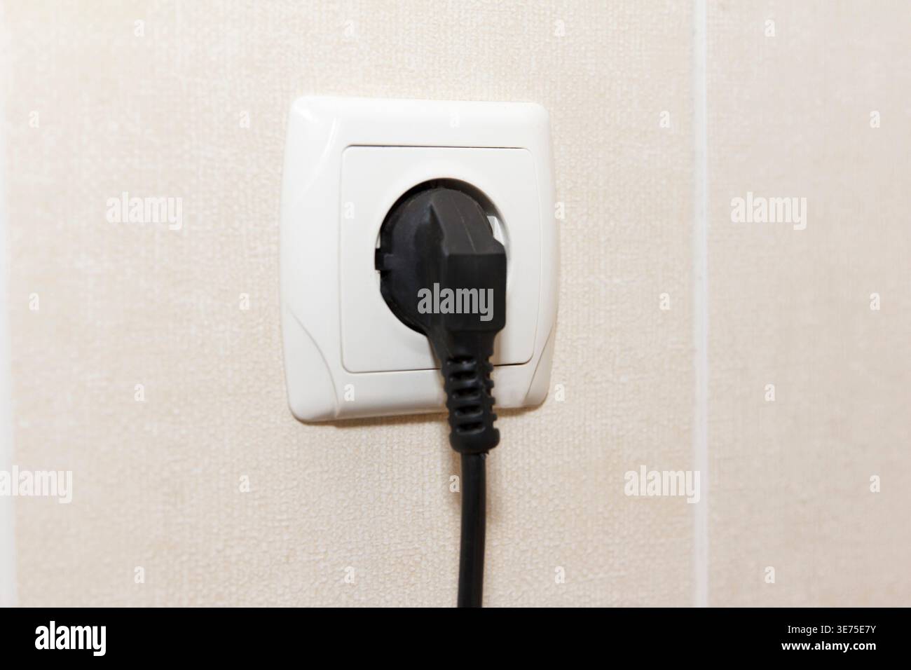 A close-up of a black electrical plug inserted into a white wall socket. Background is a light-colored wall, emphasizing plug and socket Stock Photo