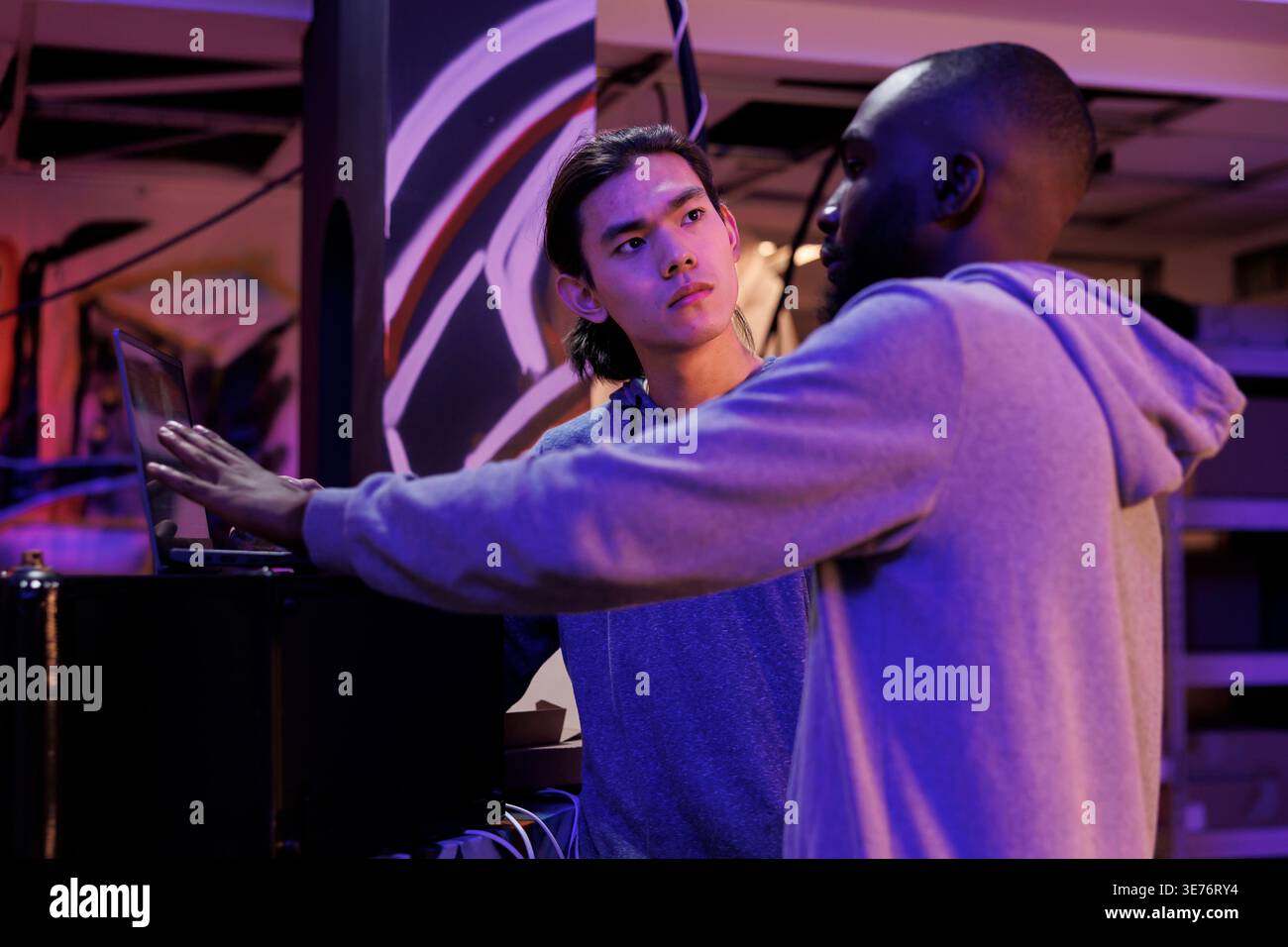 Hackers discussing malware code on laptop while planning illegal activities. African american and asian criminals talking about online server hacking technique in abandoned warehouse Stock Photo