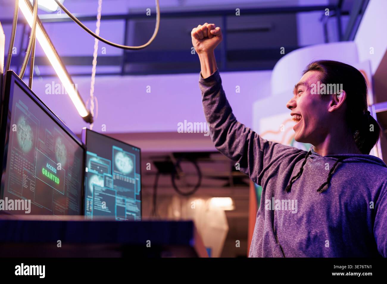 Happy hacker making victory gesture after getting server access granted. Cheerful excited asian man celebrating hacking computer network system successfully and raising fist Stock Photo