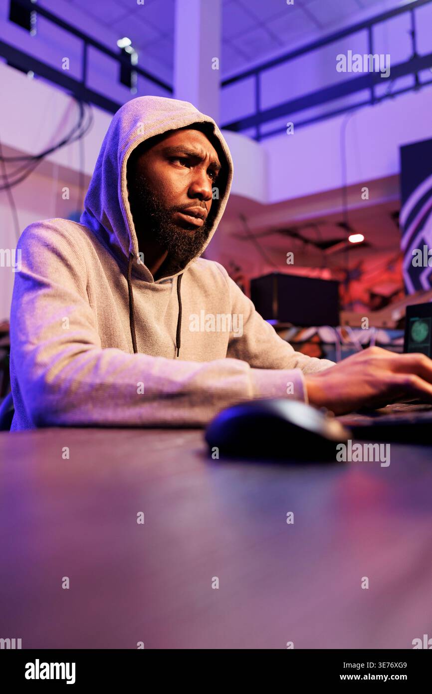 Confused african american criminal using computer for coding illegal ransomware. Concerned young hacker in hood programming malicious software to break into database server Stock Photo