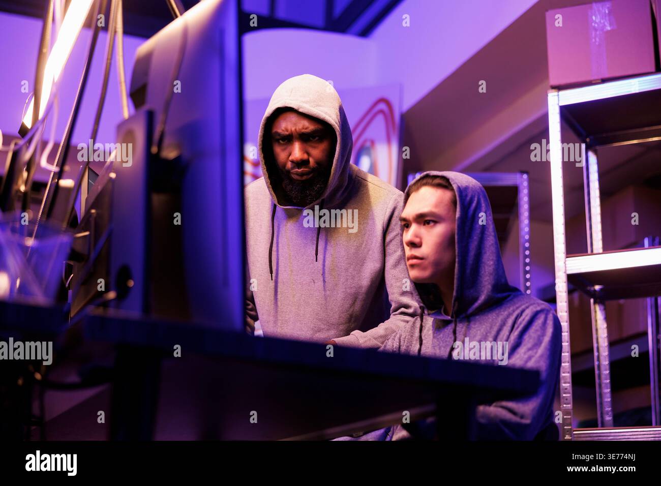 Hackers identifying vulnerabilities in system to break into database and steal data. Asian and african american criminals in hoods hacking online server and cracking password on computer Stock Photo