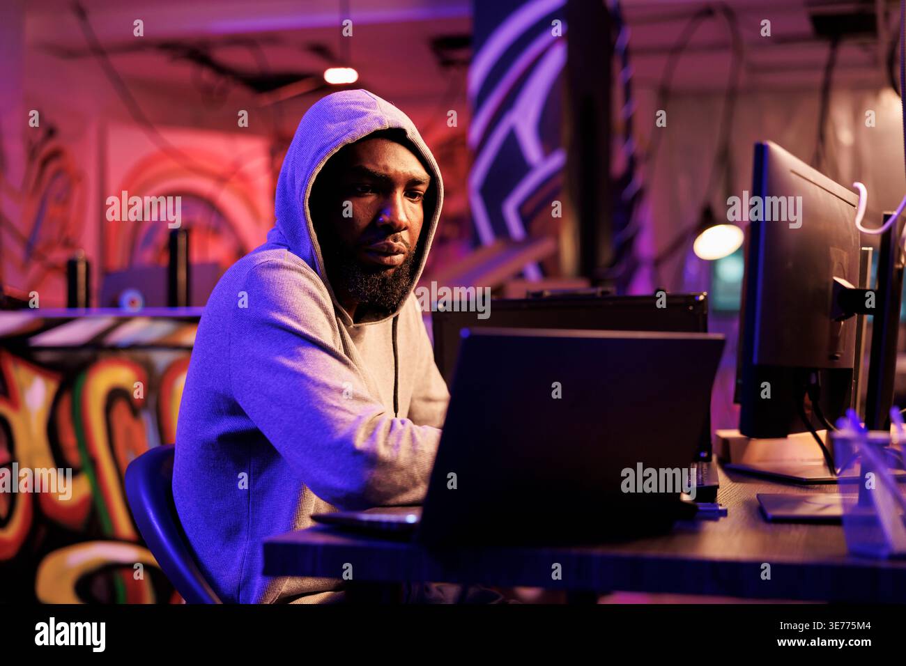 African american hacker coding computer virus to attack database. Young developer in hood cracking password and using spyware to break into online server and steal information Stock Photo