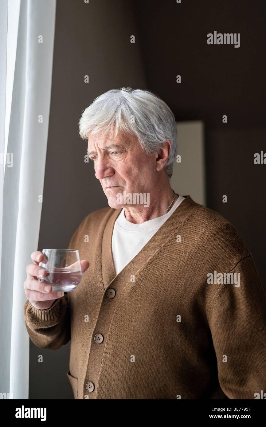 Sad sorrowful old man drinking water from glass looking out window. Loneliness and aging disease Stock Photo