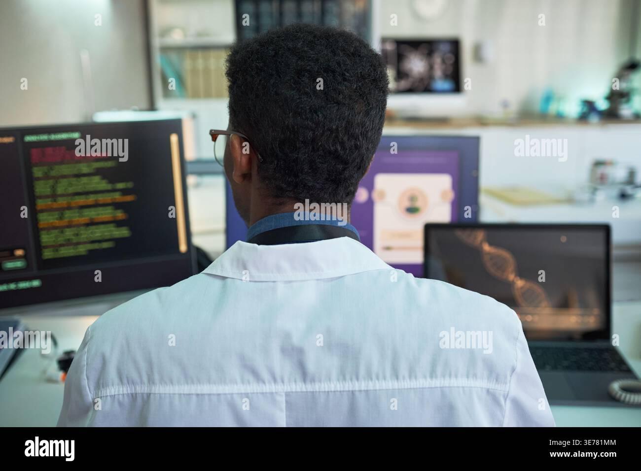Rear view of young adult Black man scientist analyzing medical data and DNA visuals on multiple screens in lab. Ideal for healthcare technology, research, diagnostics marketing Stock Photo