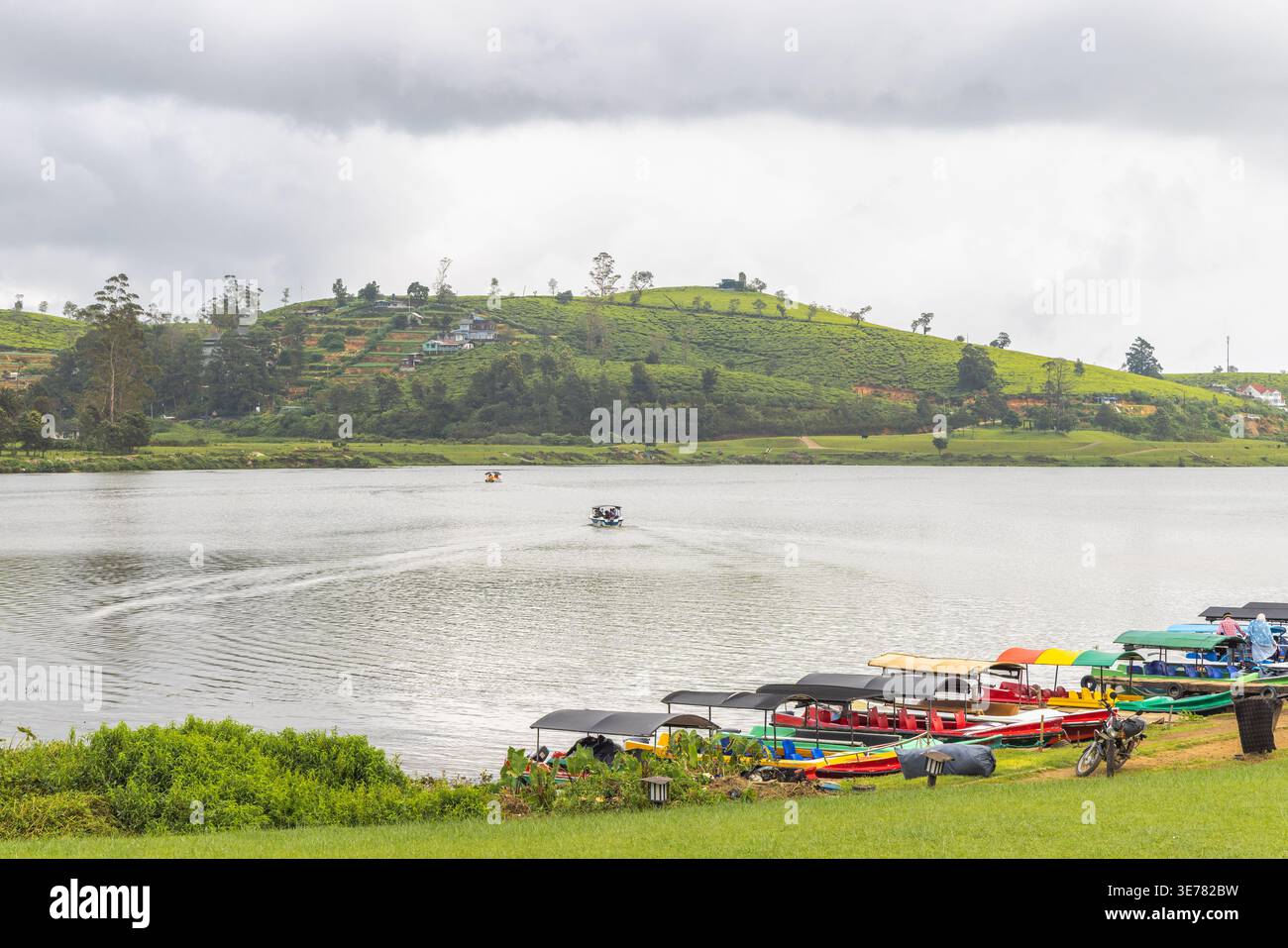 A serene lake environment with green hills, small boats docked along the shore, and a motorboat creating ripples on the water under a cloudy sky. Nuwa Stock Photo