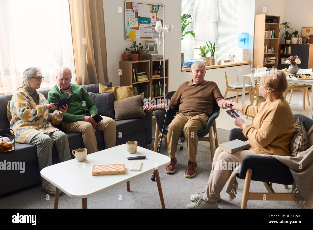 Senior men and women socializing in care home lounge, sharing photos and using smartphone during group activity. Useful for retirement living, elder care, community support marketing Stock Photo