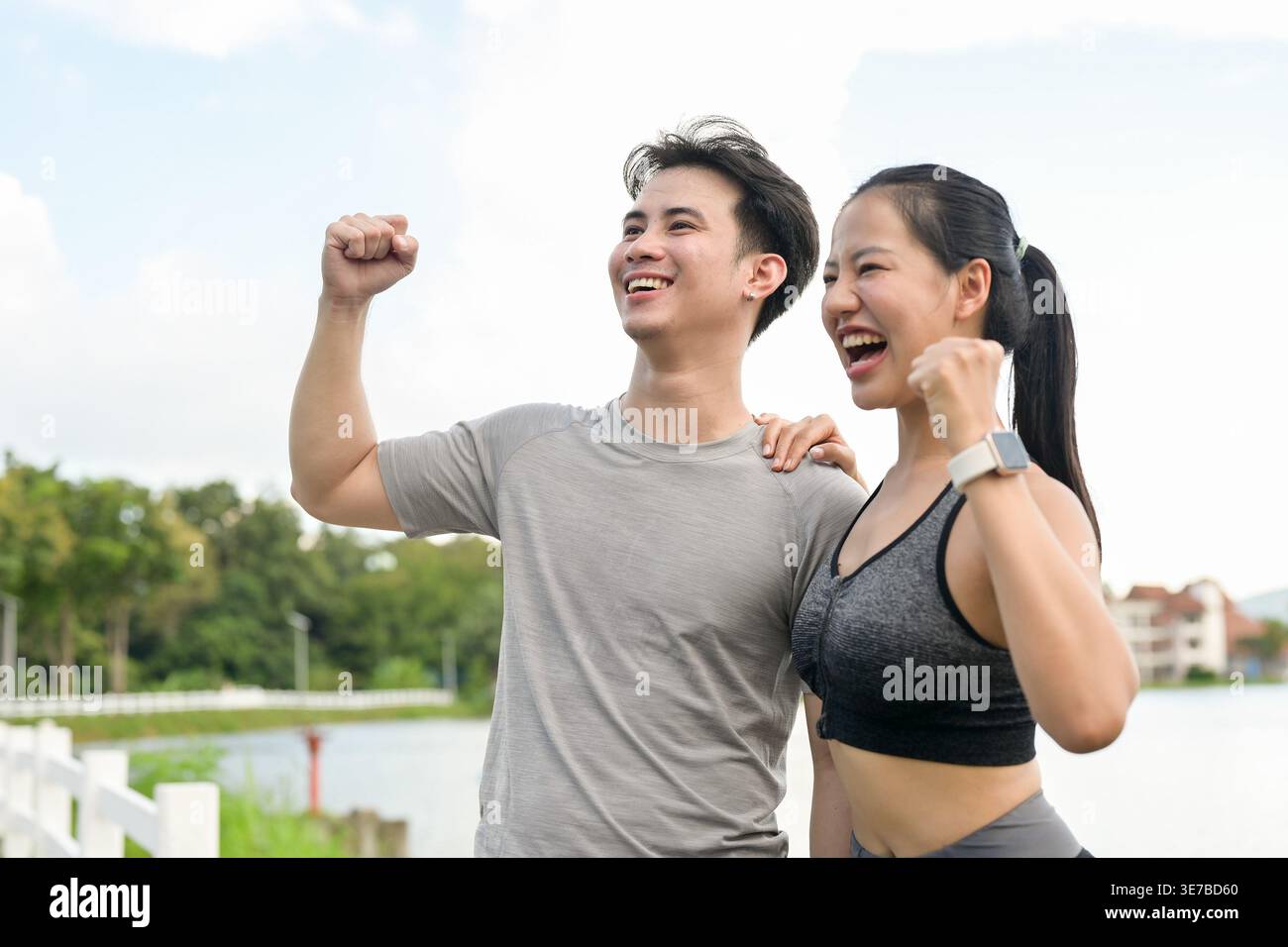 Cheerful Thai couple with raised arms and closed fists celebrating success after reaching their running target while standing together near lake in... Stock Photo