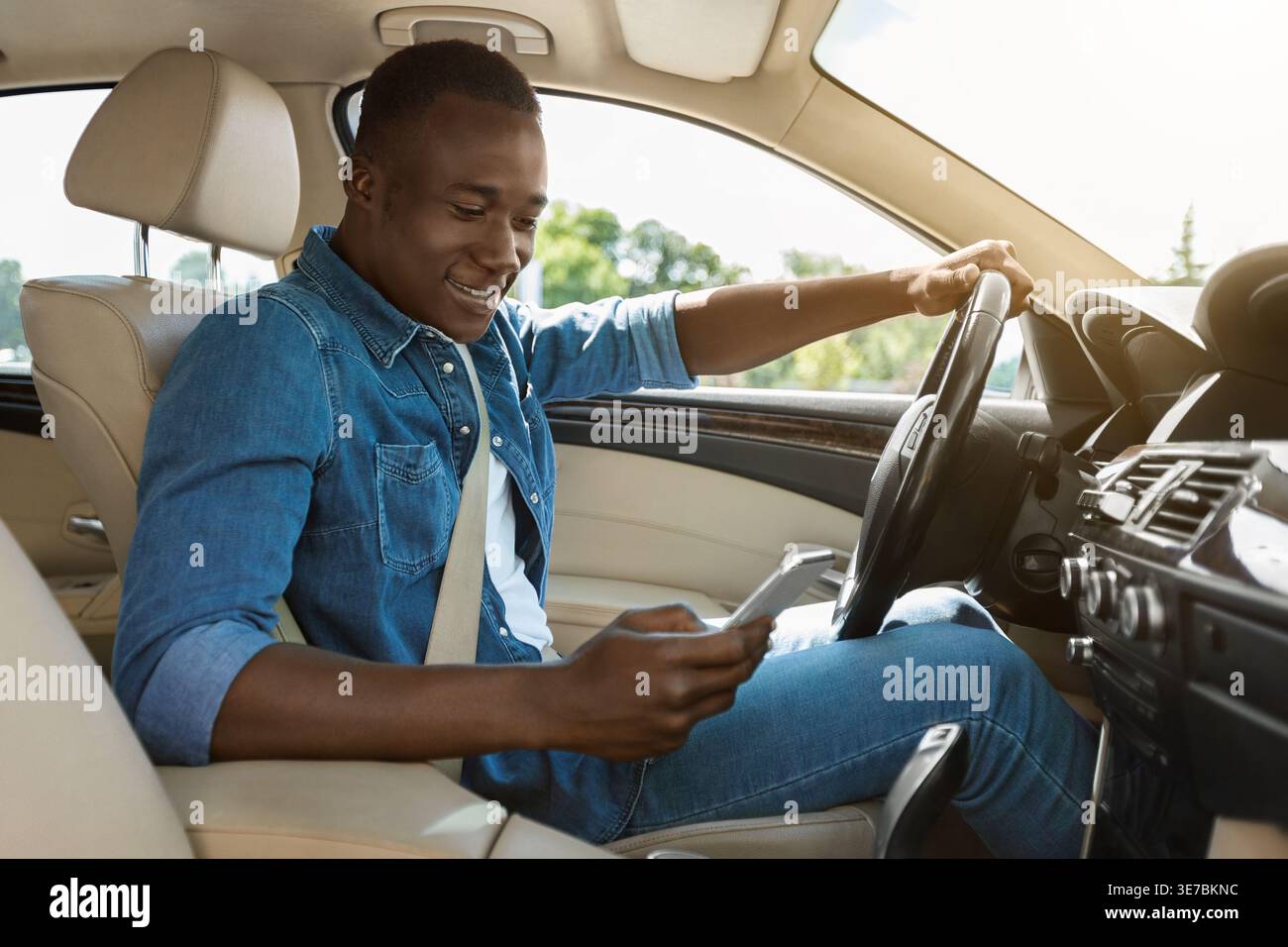 Black man wearing denim jacket using smartphone while driving car during daytime Stock Photo