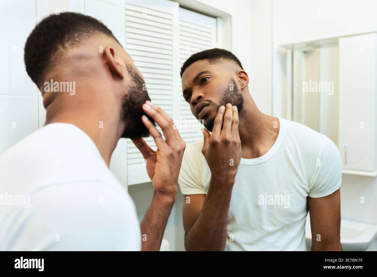 Worried Black man looking in mirror and checking his stubble standing in bathroom at home Stock Photo