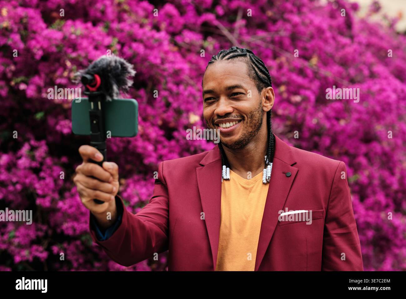 Smiling Hispanic male vlogger with braids recording video using smartphone standing in garden full of flowers during daytime Stock Photo