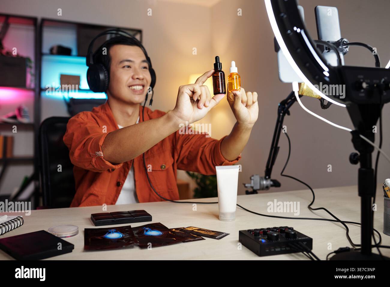 Asian man wearing headphones holding face serum bottles making content for blog while sitting at table facing ring light at home Stock Photo