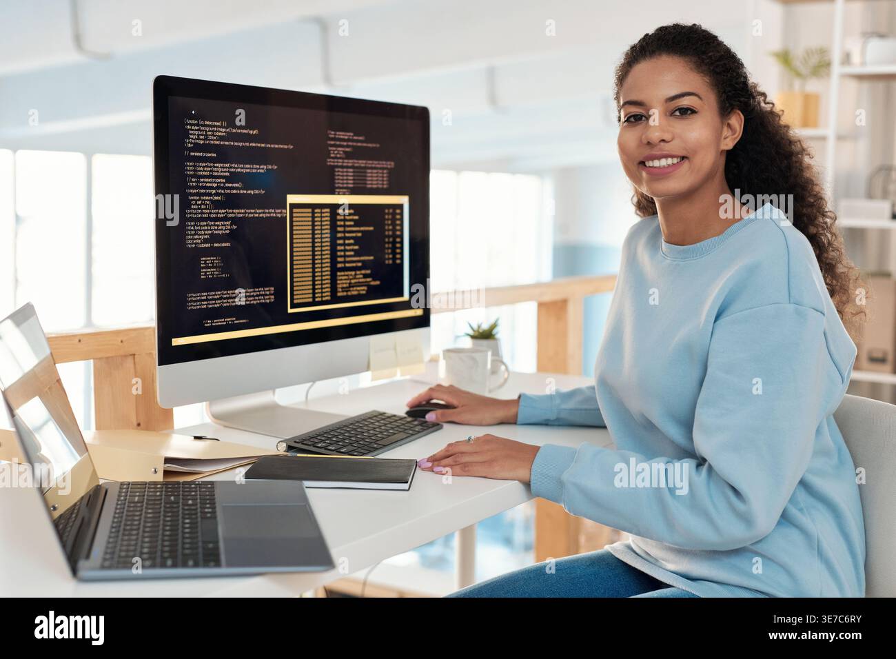 Mixed-race female programmer sitting at table with laptop and computer in office Stock Photo
