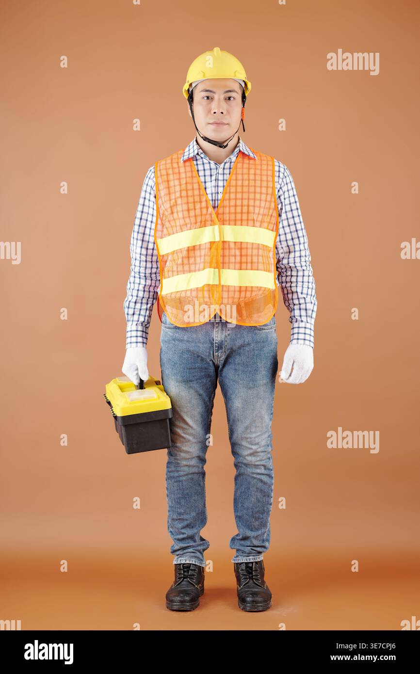 Asian male handyman wearing vest, hardhat and gloves carrying toolbox standing against brown background in studio Stock Photo