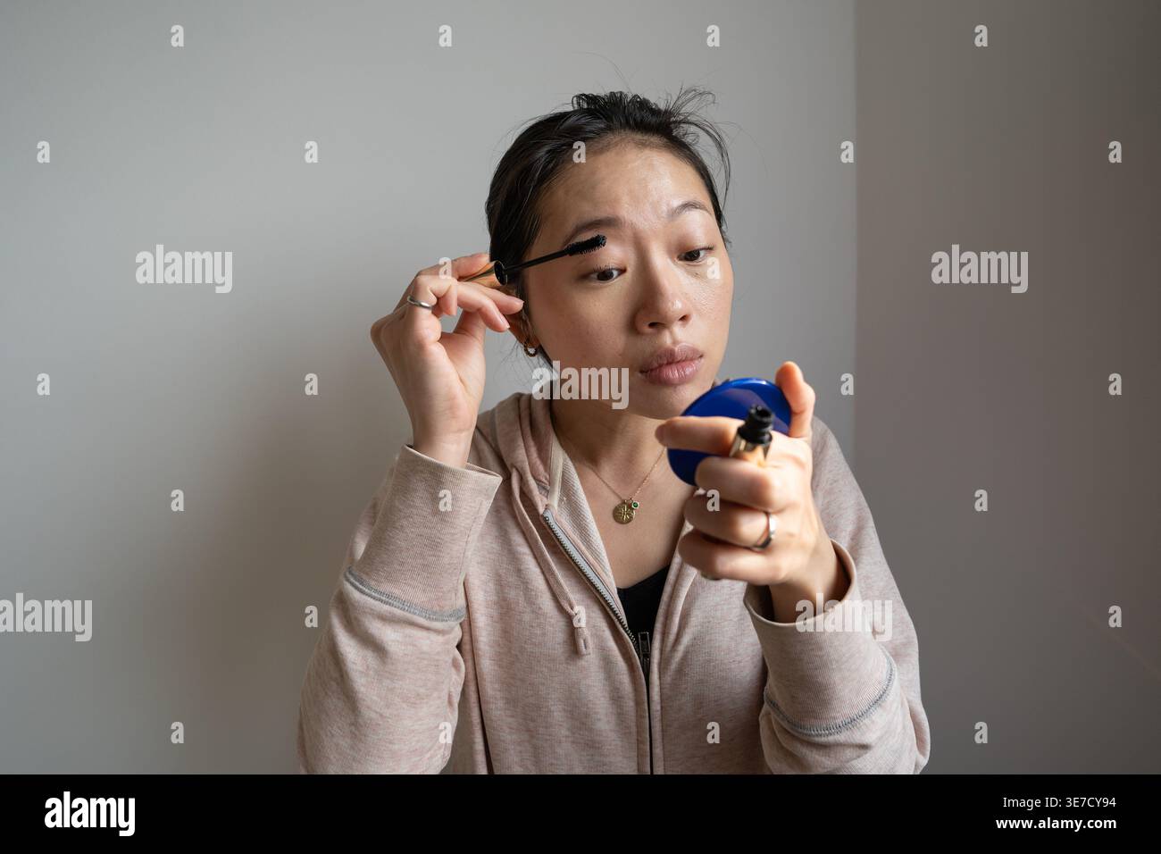 Asian woman applying mascara while looking into a handheld mirror, in a casual setting Stock Photo