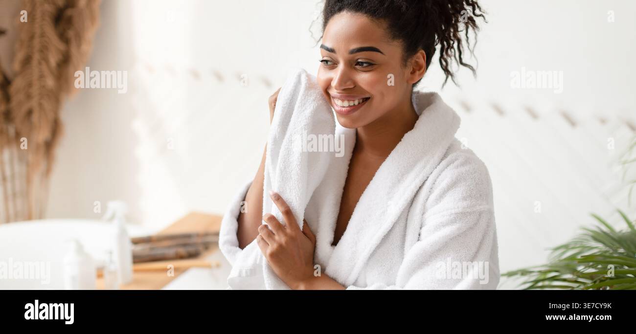 Black woman in bathrobe drying face with towel looking away while sitting in bathroom at home Stock Photo