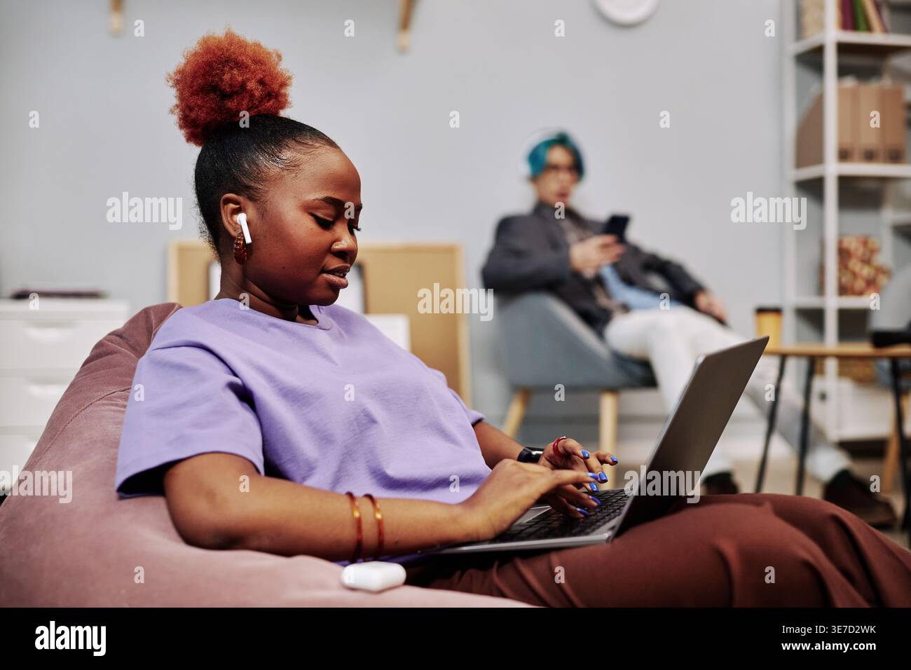 Black female programmer wearing wireless earphone working on laptop near Asian male colleague while sitting on bean bag in office Stock Photo