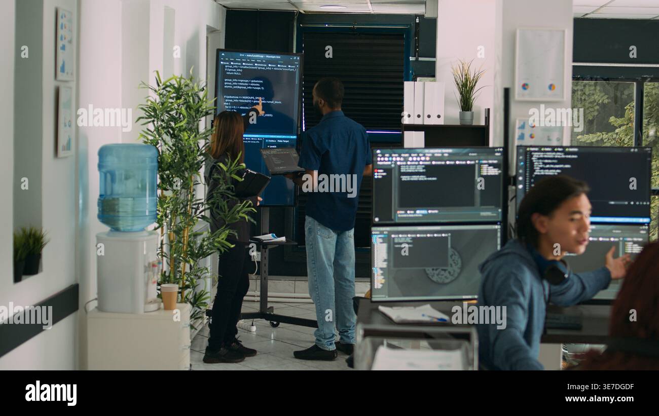 Diverse team of system developers analyzing code on big tv screen, checking errors in artificial intelligence project. Programmers working on machine learning software, developing new interface. Stock Photo