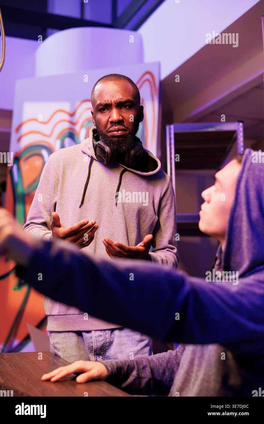 Hacker in hood pointing at illegal spyware code and asking developer for advice. Criminal showing crime partner malicious software on computer screen while cracking server password Stock Photo
