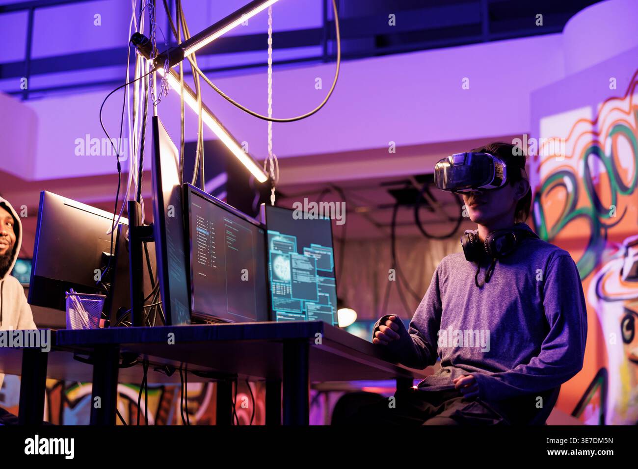 Hacker wearing vr glasses, immersed in simulation while doing illegal activity. Young criminal using virtual reality headset to code malicious software in metaverse in abandoned warehouse Stock Photo